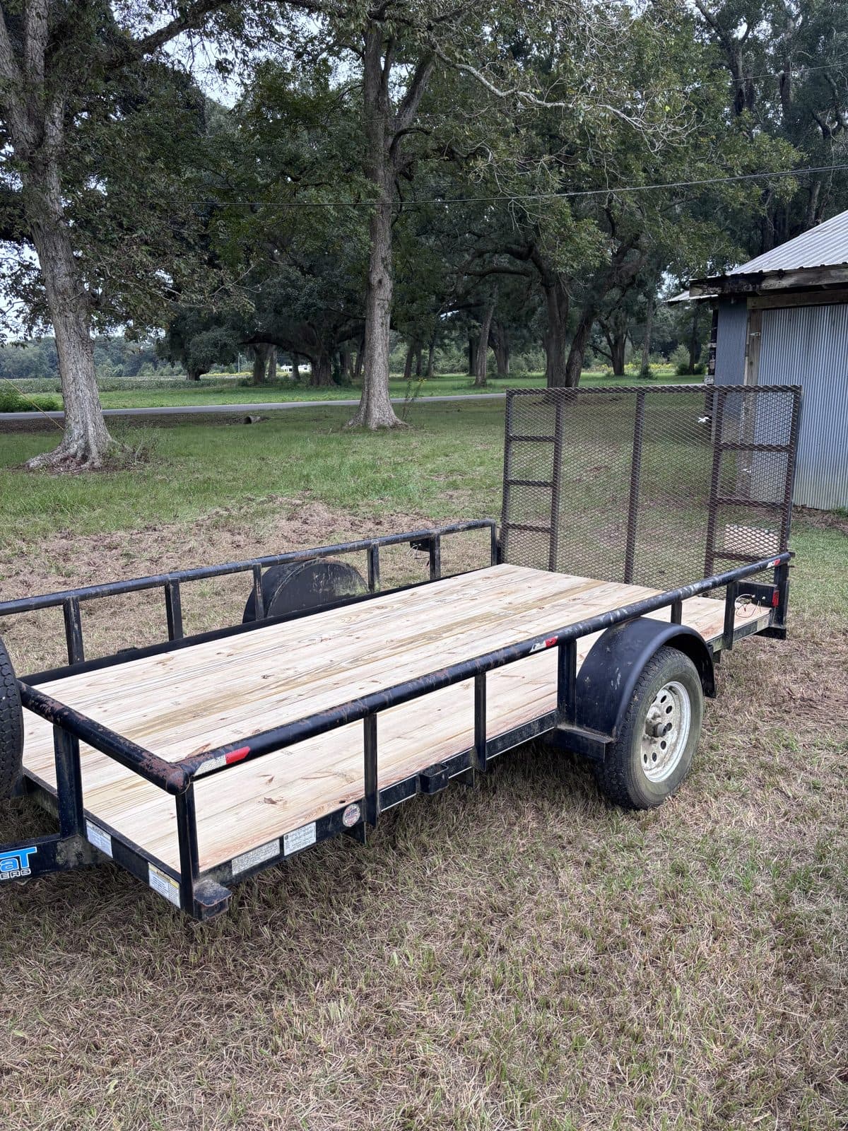 Three-quarter side view of the 6 × 12 utility trailer with the ramp raised and trees behind