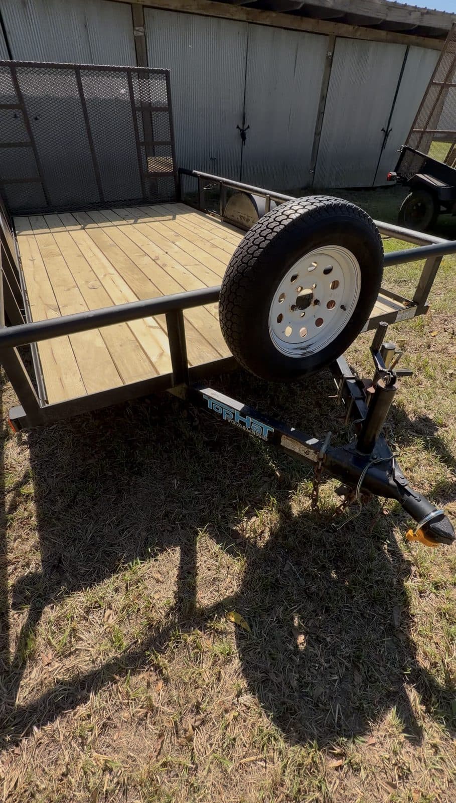 Angled view down the tongue of the second 6 × 12 utility trailer, spare tire on the rail, wood deck, and mesh ramp gate